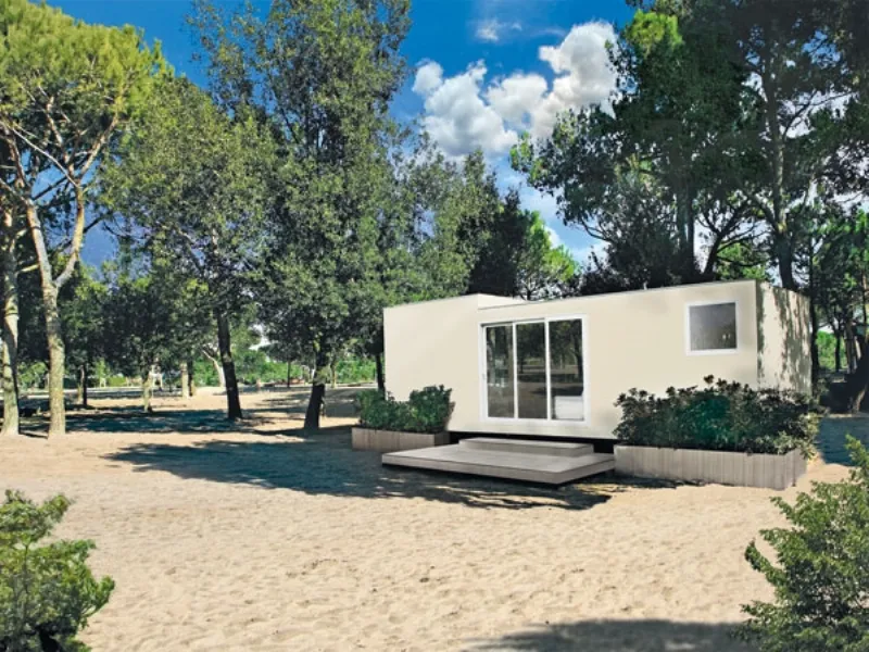 unità abitativa mobile color panna con porta finestra vetrata, fioriere con siepi in pineta con terreno sabbioso