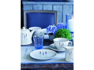 servizio da tè con teiera bianco con decorazione di lavanda, bicchiere in vetro blu sfaccettato, fiori blu e porta candela trasparente blu su tavolo in legno