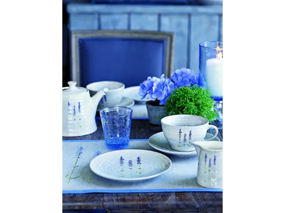 servizio da tè con teiera bianco con decorazione di lavanda, bicchiere in vetro blu sfaccettato, fiori blu e porta candela trasparente blu su tavolo in legno