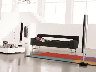 divano a tre posti nero con gambe in alluminio inserito in spazio living bianco con tappeto geometrico multicolore e casse verticali