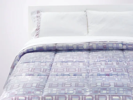 letto matrimoniale con biancheria bianca e copriletto con logo dai toni pastello lavanda e lilla